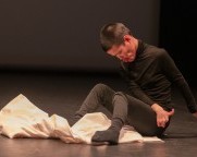 Eine zierliche Person mit kurzen schwarzen Haaren, die schwarze Kleidung trägt, sitzt auf einer Bühne und lehnt sich leicht nach vorne. Ihre Arme sind über beide Oberschenkel nach links ausgestreckt. Ein weisses Tuch liegt unter den Füssen.