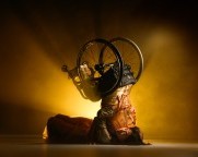 Ein männlicher Rollstuhltänzer und eine ostasiatische Tänzerin liegen auf dem Boden in einer Umarmung. Die Tänzerin liegt auf dem Rücken, und der Rollstuhltänzer ist senkrecht zur anderen Tänzerin, aber kopfüber, als ob er einen Kopfstand macht.