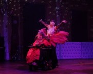 Eine Person in einem roten Kostüm ist im Rollstuhl. Am Rollstuhl sind lange roten Federn befestigt. Hinter der Rollstuhltänzerin steht eine Person mit Rollschuhen.  Sie hat die Arm ausgestreckt und trägt glamouröse Kleidung und eine Stoffblume im Haar. 