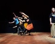 Eine Person steht am rechten Bühnenrand. Eine Gruppe von Tänzern, eine Tänzerin ist im Rollstuhl, bewegt sich in der Bühnenmitte. Die Arme sind ausgestreckt und die Körper neigen sich nach links.