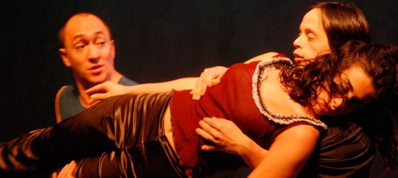 Bühnenaufnahme. Zwei Männer halten eine Frau von hinten gemeinsam in einer schwebenden Liegeposition. Ihr Augen sind geschlossen.