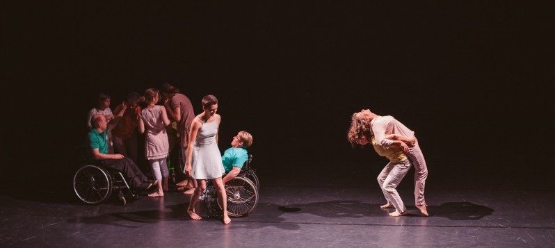 Acht Personen, zwei davon im Rollstuhl sind am Tanzen, ein einziges Licht beleuchtet die Szene.