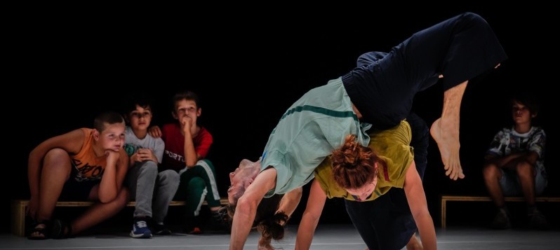 Bühnenaufnahme des Stückes «Equality», Von links schlägt ein grossgewachsener, Mann rückwärts eine Brücke über die Schultern einer kleineren Frau in Liegestütz. Beide haben ihre langen Haare zu einem losen Knoten hochgesteckt. 