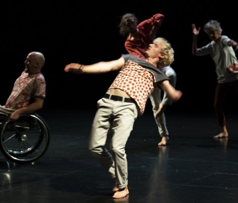 Ein Quartett mit drei Männern und einer Frau unterschiedlichen Alters tanzt dynamisch verteilt im schwarzen Bühnenraum. Alle tragen unterschiedliche Oberteile und Hosen in Rot- und Grautönen. 