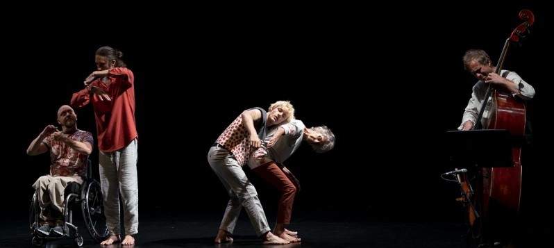 Hell beleuchtet stehen vier Tanzende und ein Kontrabassist auf einer Linie im sonst schwarzen Bühnenraum. Der Musiker spielt am rechten Rand. Je zu zweit tanzen weiter links drei Männer und eine Frau unterschiedlichen Alters - stehend und ein Mann sitzend