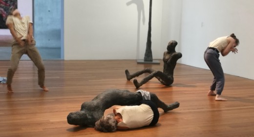 Ein Tanztrio mit hellen T-Shirts, dunkeln oder beigen Hosen durchmischt sich in einem modernen Galerieraum mit sitzend, stehend und liegenden menschlichen Skulpturen, in dunkler Bronze. Das Trio teilt und ergänzt die kräftigen, gebogenen Formen der Statue