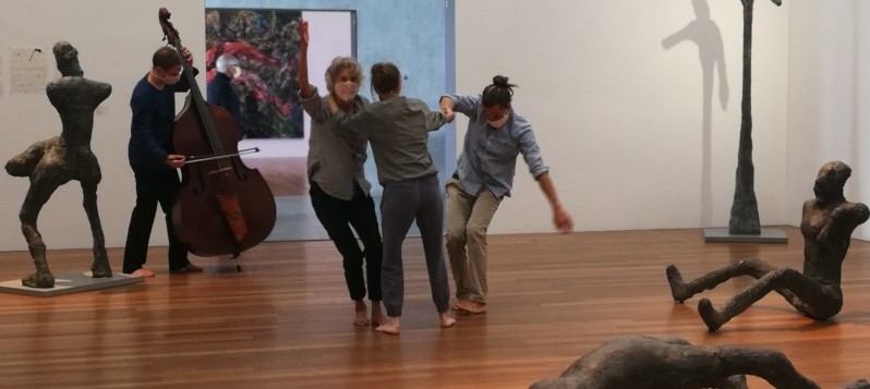 In der Mitte eines modernen Galerieraumes halten sich zwei Frauen und ein Mann voneinander weglehnend momentan im Fallen auf. Links hinter ihnen spielt ein Kontrabassist. Um sie verteilt sind vier liegende, sitzende oder stehende menschliche Skulpturen, k