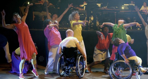 Die BewegGrund Performance Gruppe tanzt ausgelassen vor einer erhöhten Bühne, wo das Tabula Musica Orchester spielt.