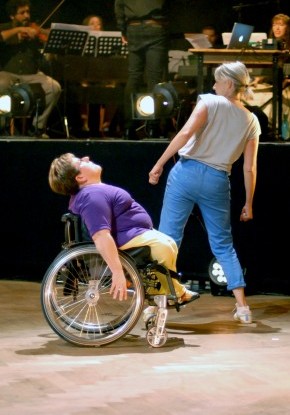 Zwei Frauen tanzen leicht versetzt nebeneinander vor einer erhöhten Bühne, auf der ein Orchester spielt. Die Tänzerinnen, eine links im Rollstuhl sitzend, die andere breit stehend, lehnen voneinander weg.
