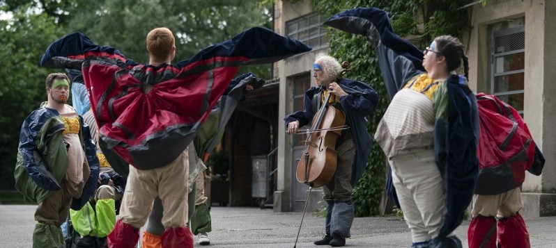 Auf einer Terrasse spielt ein Cellist zwischen acht diversen Tänzer:innen in flügelartigen Capeanzügen.