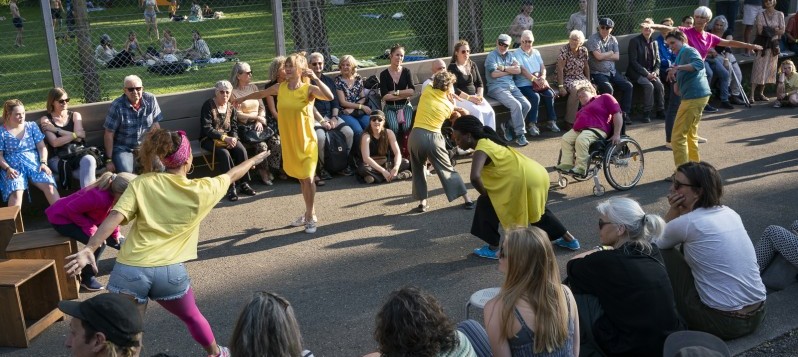 Acht diverse Tänzer:innen bewegen sich ringsum umgeben vom sitzenden Publikum über die Länge eines mit Netzen abgeteilten Sportplatzes. Dahinter liegt eine Parkwiese mit Bäumen.