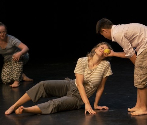 Bühnenaufnahme des Stückes Milles Sens. Ein jugendlicher Tänzer mit Trisomie 21 bückt sich und hält einer seitlich sitzenden jungen Tänzerin eine Zitrone entgegen. Sich sinnlich zuwendend riecht diese genüsslich daran.