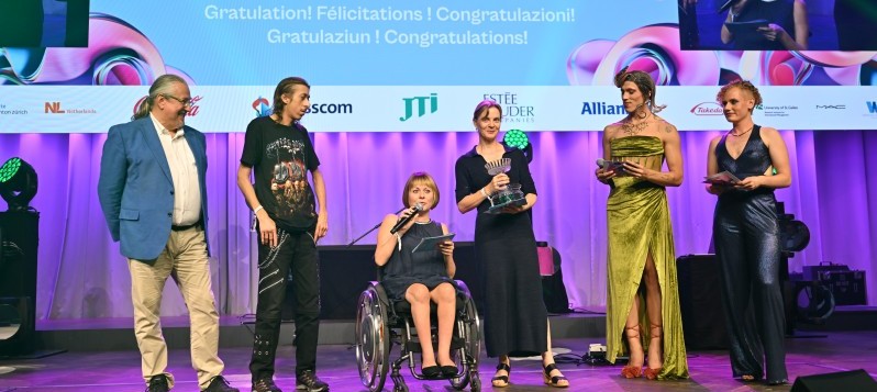 Mirjam Gasser, die Präsidentin von BewegGrund bedankt sich bei den Veranstaltern und dem Publikum. Neben ihrem Rollstuhl hält Susanne Schneider die gläserne Trophäe. Vier weitere Personen, zwei davon gender-fluid, stehen bei ihnen. 