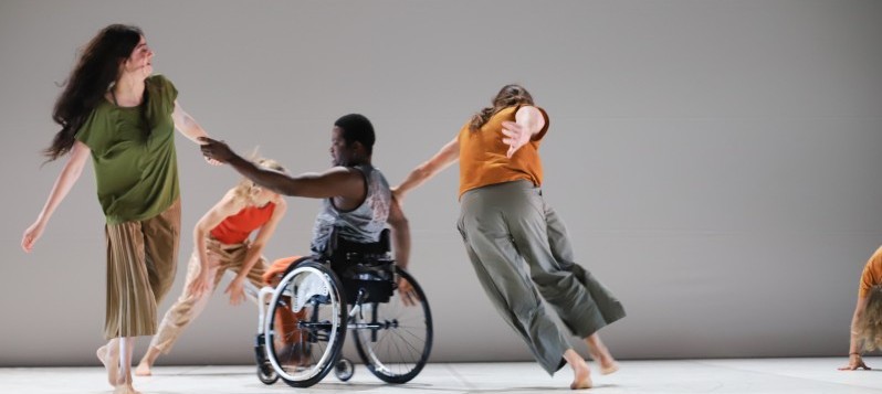 Im weissen Bühnenraum hält ein Tänzer, der in einem Rollstuhl sitzt, eine Tänzerin mit ausgestrecktem Arm an der Hand fest. Ein dritter Tänzer hält sich an der Schulter des Tänzers im Rollstuhl. Das Trio dreht sich im Kreis.