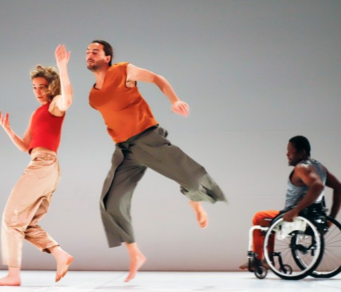 Eine Tänzerin hält im Laufen ihren Unterarm angewinkelt nach oben. Hinter ihr scheint ein Tänzer im Sprung über ihre Schulter zu gucken. Ein weiterer Tänzer fährt hinten im Rollstuhl quer durch den weissen Bühnenraum. 