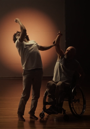 Videostill von Tanz ist eine Sprache. Ein schwarzer Mann mit dichtgekraustem Haar umschliesst mit seiner Hand sanft den Ellbogen eines weissen Mannes im Rollstuhl. Dieser führt ihn mit gehobenem Arm in eine langgestreckte Rückbeuge.
