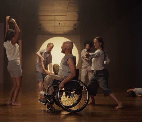 Videostill. Mehrere Menschen tanzen in einem breiten Gang vor einer dunklen Leinwand mit einem weissen Lichtkreis. Statuenhaft verharren sie mitten in der Bewegung: gestreckt, gebogen, sitzend in einem Rollstuhl oder am Boden liegend.