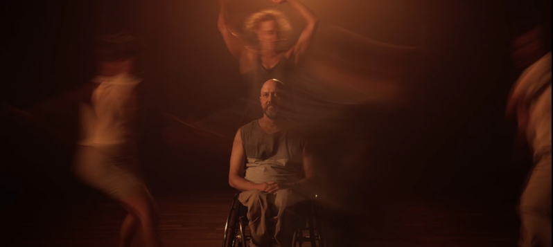 Videostill von Tanz ist eine Sprache. Ein weisser Mann mit Glatze und graumeliertem Bart sitzt still im Rollstuhl und schaut entspannt direkt nach vorne. Lebhaft, nebelgleich verwischt umtanzen ihn drei Personen in weichem, orangem Licht.