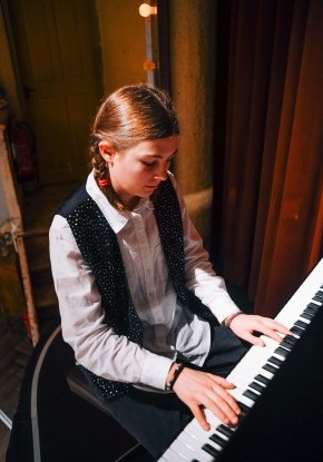 Eine junge Frau mit Mittelscheitel und Zöpfen sitzt hoch konzentriert am Klavier und spielt. Sie trägt ein weisses Kragenhemd, darüber ein schwarzes Gillet mit feinen Glitzertupfen.