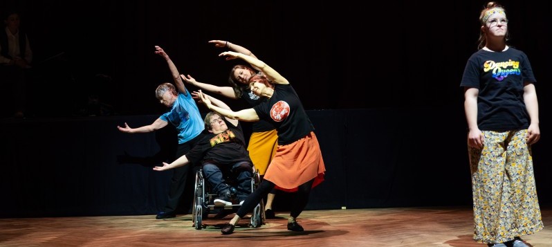 Allein steht eine junge Frau mit Trisomie 21 vorne rechts auf der Bühne. Auf ihrem T-Shirt steht Dancing Queen geschrieben. Drei stehende Frauen und eine Frau im Rollstuhl wiegen im Hintergrund ihre Körper mit gehobenen Armen zur Seite.