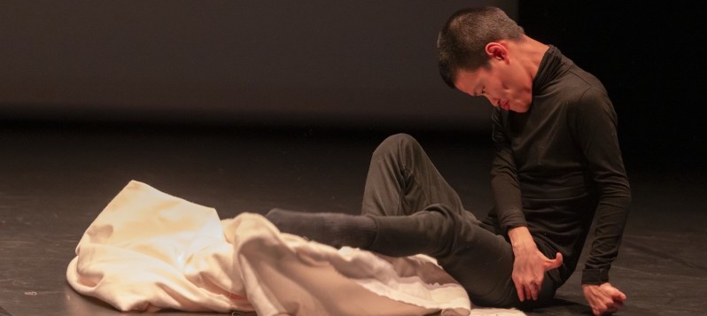 Eine zierliche Person mit kurzen schwarzen Haaren, die schwarze Kleidung trägt, sitzt auf einer Bühne und lehnt sich leicht nach vorne. Ihre Arme sind über beide Oberschenkel nach links ausgestreckt, und ein weisses Tuch liegt unter den Füssen. 