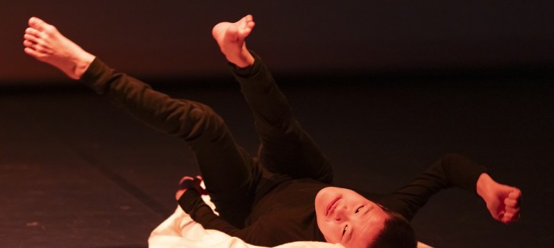 Eine Person mit kurzen schwarzen Haaren und zierlichem Körper in schwarzer Kleidung liegt auf einer Bühne, umgeben von einem weissen Stoff. Die Beine sind in die Luft gestreckt, während die Arme nach unten zeigen. 