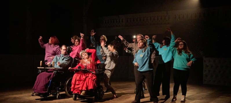 Eine gemischte Gruppe von zehn Personen bewegen ihre Oberkörper in der Bühnenmitte. Acht Personen stehen nebeneinander, ihre Arme wechselnd nach oben schwingend. Zwei Personen sind im Rollstuhl auch sie bewegen gemeinsam mit der Gruppe ihren Oberkörper. 