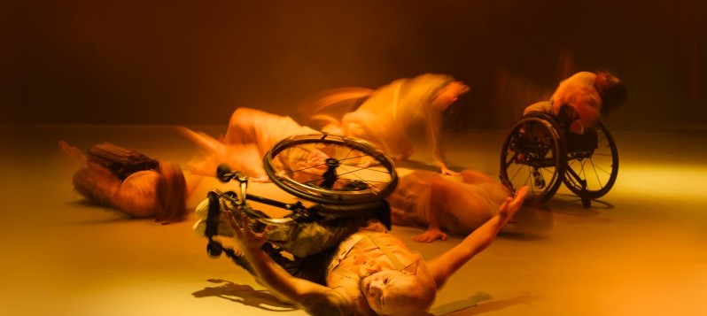 Das Bild hat eine goldene Farbe mit atmosphärischem Dunst und schwarzem Hintergrund. Im Vordergrund liegt eine Rollstuhlfahrerin nach links gerichtet auf dem Boden. Ihre Arme sind zur Seite ausgestreckt. Sie blickt nach links. 