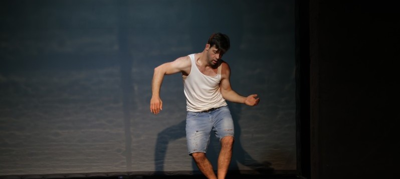 Ein Mann tanzt in der Mitte der Bühne. Die Knie sind leicht gebeugt. Beide Arme angewinkelt. Der rechte Arm schwingt leicht nach vorne, der linke Arm nach hinten. Der Tänzer blickt nach unten zu seinem linken Fuss. 