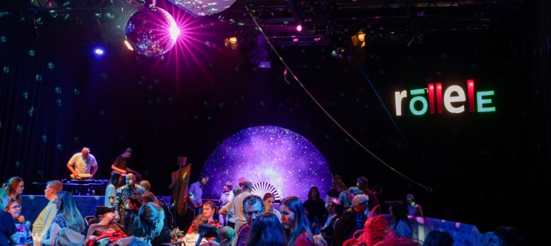 Viele fröhliche, tanzende Menschen mit Rollschuhen oder im Rollstuhl auf der Discofläche. Etwas erhöht steht ein DJ an einem in DJ-Pult. An der Decke strahlt eine Discokugel und im Hintergrund leuchtet in Leuchtschrift "röllele".  