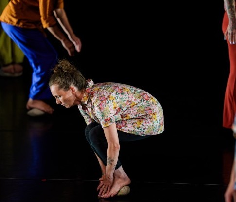 Eine Gruppe von Tänzer*innen ist auf der Bühne. Im Zentrum ist eine Tänzerin mit hochgesteckten braunen Haaren, einer kurzen Bluse mit Blumen und dunklen Hosen. Sie balanciert auf einem  Stein.