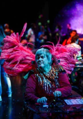 Eine Person in einem roten, festlichen  Kostüm sitzt im Rollstuhl. Am Rollstuhl sind lange roten Federn befestigt. Im Hintergrund, leicht verschwommen, tanzen Menschen.