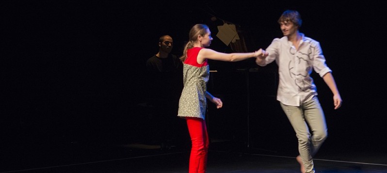 Bühnenaufnahme. Eine junge, feingliedrige Frau hält die Hand eines jungen Mannes über Kreuz. Sie steht und er geht in einem Bogen um sie.