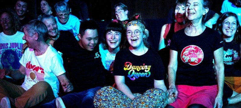 Eine fröhliche Gruppe von Menschen sitzt eng beieinander auf dem Boden, lacht und geniesst den Moment. fdrucken wie „Dancing Queen“ – vermitteln Lebensfreude, Gemeinschaft und Inklusion.