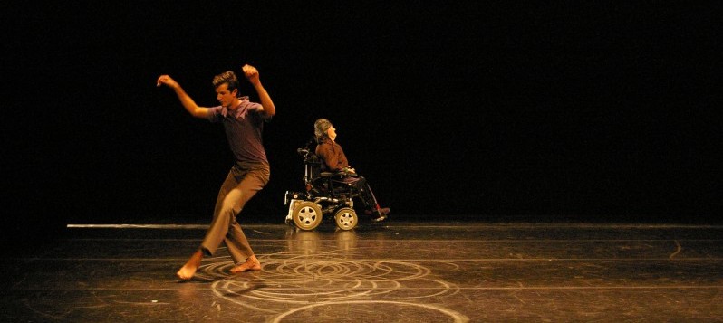Ein Tänzer im Rollstuhl und ein Tänzer sind zusammen auf der Bühne, auf dem Boden sind runde Kreidekreise.