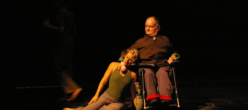 Eine Tänzerin sitzt auf dem Boden und lehnt sich an den Tänzer im Rollstuhl.