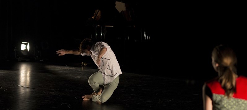 Bühnenaufnahme. Ein junger Mann in einem weissen Hemd balanciert in einer tiefen Spirale. Im Vordergrund schaut ihm eine sitzende Frau zu.