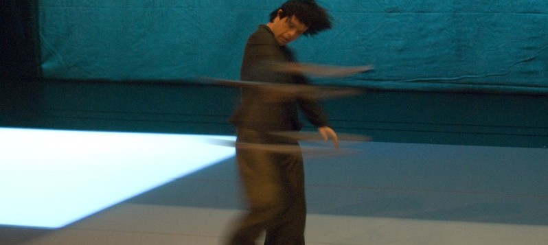 Bühnenaufnahme. Ein kleiner Mann in dunkler Kleidung dreht sich im Kreis nach rechts. Sein Arm verschwimmt in Bewegung.