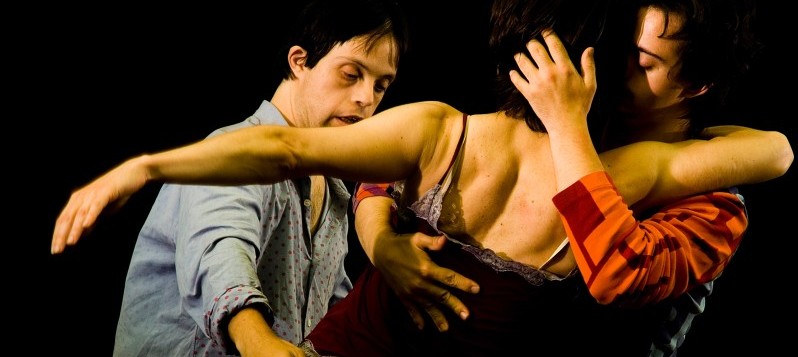 Bühnenaufnahme. Rechts umarmt ein Mann eine Frau. Ihre Köpfe sind sich innig nah. Ein Mann mit Trisomie 21 umrahmt das Paar von links.