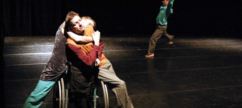 Im Vordergrund umarmen sich zwei Tanzende, ein weiterer Tänzer tanzt im Hintergrund solo.