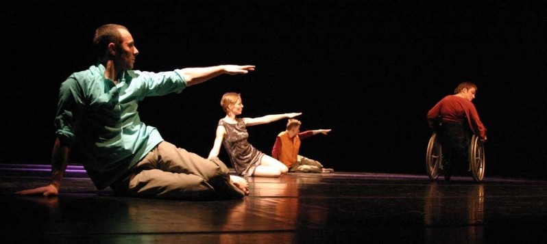 Drei Tänzerinnen und Tänzer sitzen auf dem Boden und zeigen mit ausgestreckten Händen auf eine Rollstuhltänzerin.