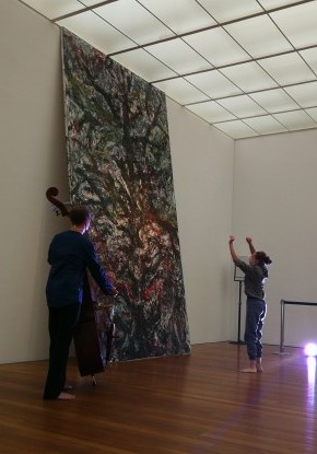 In der Mitte eines modernen Galerieraumes steht eine Tänzerin mit im Laufe gebogenen Knien und wringt ihren Körper mit asymmetrisch angewinkelten Armen nach hinten. Links hinten spielt ein Kontrabassist. Um sie verteilt sind vier liegende, sitzende oder s