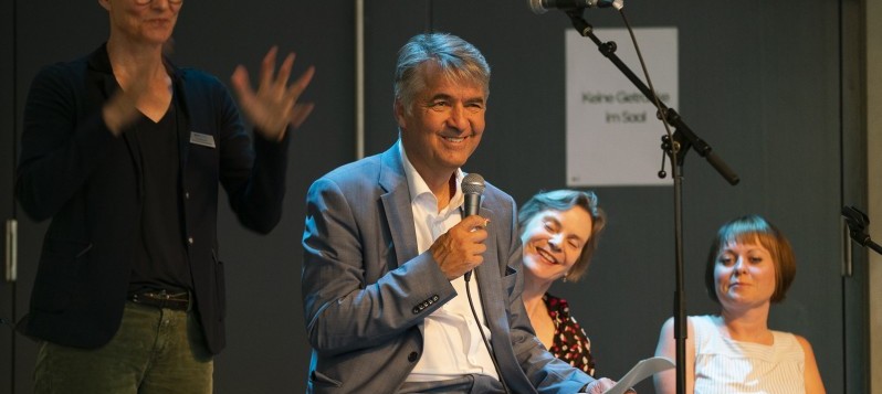 Auf einer Terrasse spielt ein Cellist zwischen acht diversen Tänzer:innen in flügelartigen Capeanzügen.