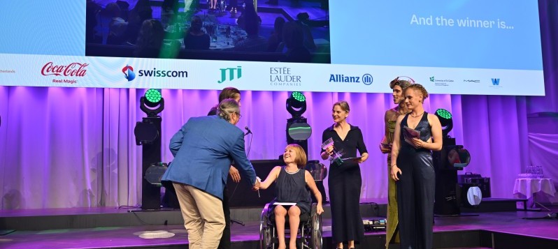 Mirjam Gasser, die Präsidentin von BewegGrund bedankt sich bei den Veranstaltern und dem Publikum. Neben ihrem Rollstuhl hält Susanne Schneider die gläserne Trophäe. Vier weitere Personen, zwei davon gender-fluid, stehen bei ihnen. 