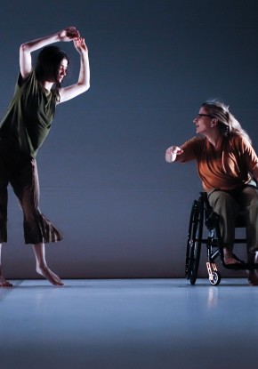 Eine Tänzerin hält im Laufen ihren Unterarm angewinkelt nach oben. Hinter ihr scheint ein Tänzer im Sprung über ihre Schulter zu gucken. Ein weiterer Tänzer fährt hinten im Rollstuhl quer durch den weissen Bühnenraum. 