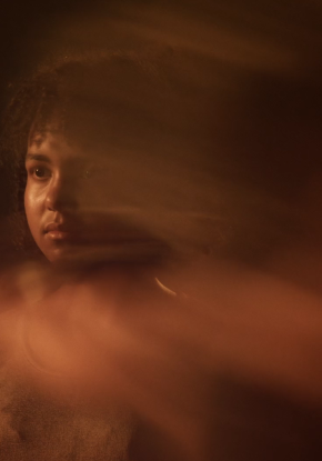 Videostill. Mehrere Menschen tanzen in einem breiten Gang vor einer dunklen Leinwand mit einem weissen Lichtkreis. Statuenhaft verharren sie mitten in der Bewegung: gestreckt, gebogen, sitzend in einem Rollstuhl oder am Boden liegend.