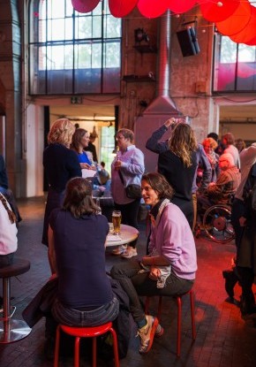 Eine Gruppe von Tänzer*innen ist auf der Bühne. Im Zentrum ist eine Tänzerin mit hochgesteckten braunen Haaren, einer kurzen Bluse mit Blumen und dunklen Hosen. Sie balanciert auf einem  Stein.