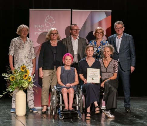 Mirjam Gasser (im Rollstuhl) und Susanne Schneider sitzen nebeneinander und halten gemeinsam die Urkunde des Bürgi-Willert-Stiftung Preises.